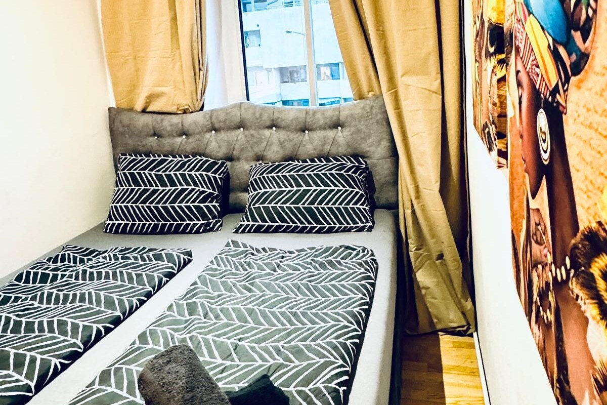 Ein Bett mit gemusterten Textilien, ein Fenster mit Vorhängen und eine Wand mit einem farbenfrohen Wandbild.