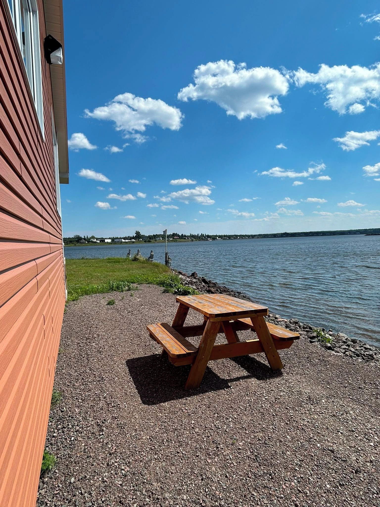 Una mesa de picnic de madera sobre un sendero de grava junto a un cuerpo de agua, con un edificio a la izquierda y un cielo despejado.