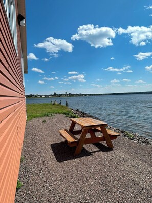 Una mesa de picnic de madera sobre un sendero de grava junto a un cuerpo de agua, con un edificio a la izquierda y un cielo despejado.