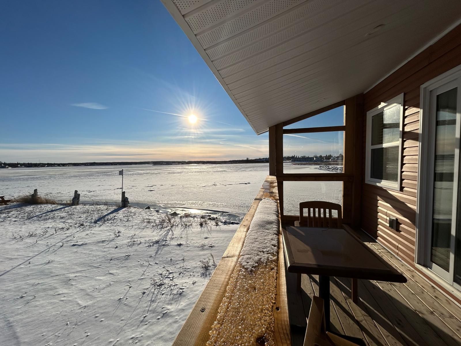 Una cabaña de madera con vistas a un lago helado, una mesa y una silla afuera.