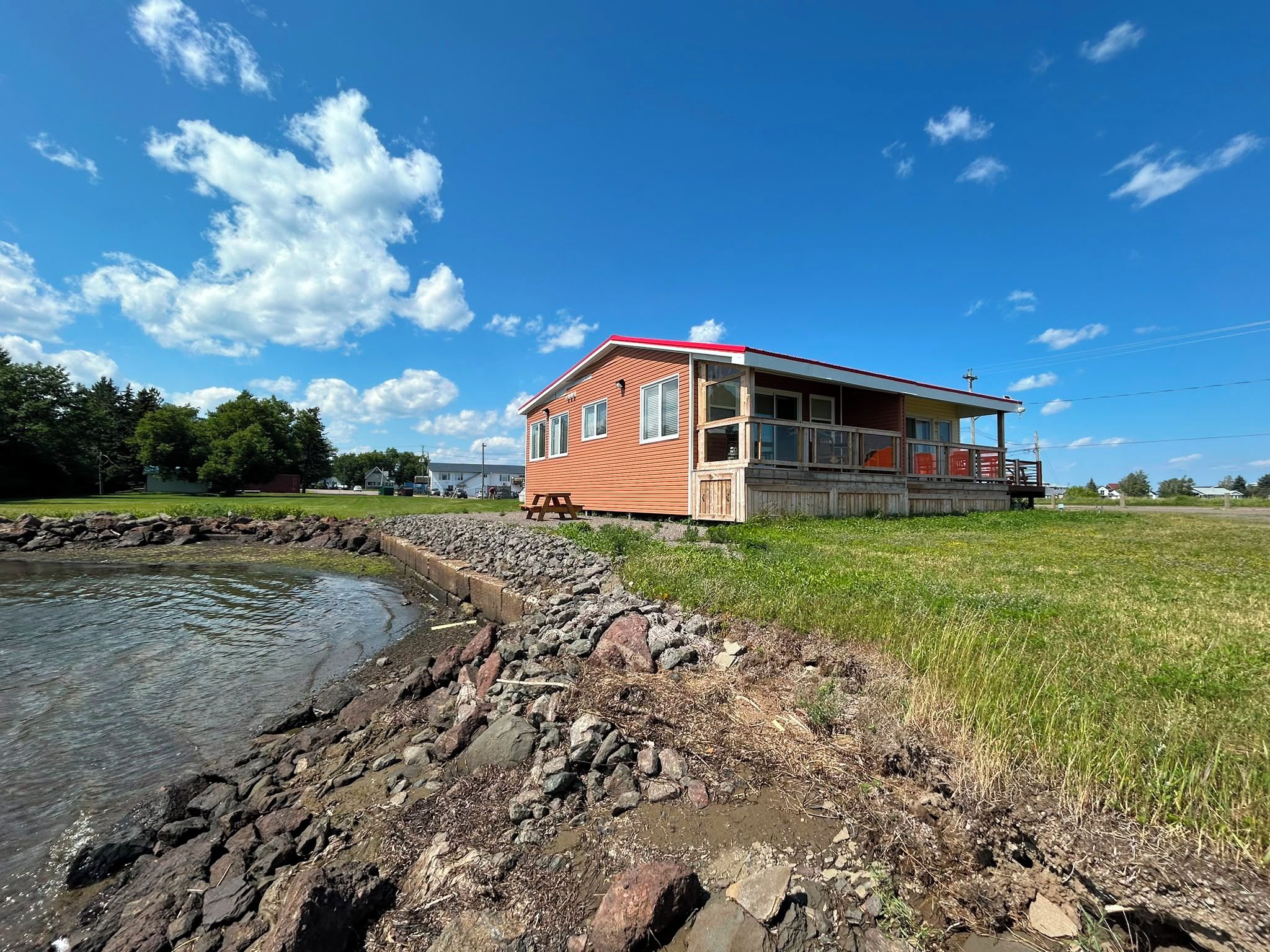 A single-story building with a red roof, a covered porch, and a small deck. It is situated next to a body of water with a rocky shoreline.