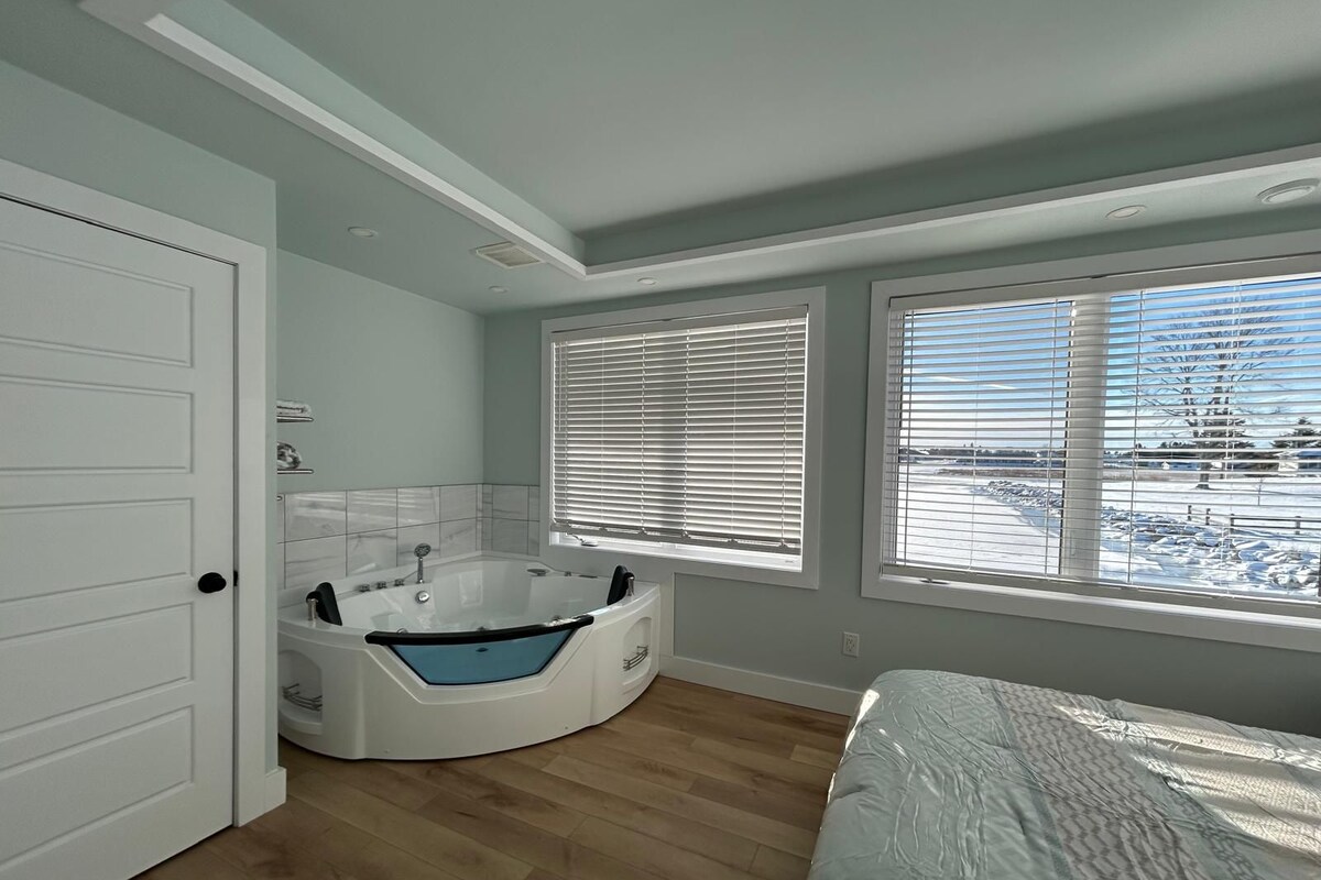 Un baño moderno con una bañera independiente, ducha y retrete. Hay ventanas amplias con persianas, y vistas a un paisaje nevado.