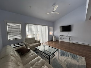 A modern living room with a sofa, a glass coffee table, a ceiling fan, and a flat-screen TV mounted on the wall.