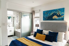 A hotel room with a bed, a painting, a bathroom with a shower, and a view through a glass door.
