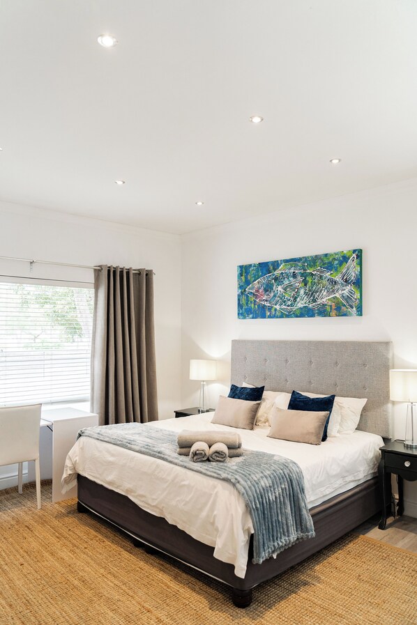 A modern bedroom with a large bed, bedside tables, a window with curtains, and a painting on the wall.