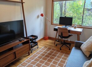 A room with a TV, a desk with a computer, a chair, a sofa, and a window with a view of trees. - White Cap Retreat: 3-Bedroom Sandy Riverfront & Mt Hood Getaway (Sandy)