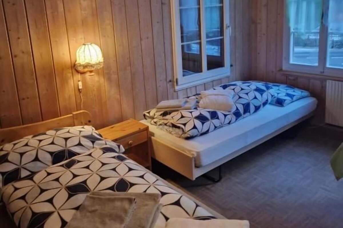 A wooden cabin room with two beds, a window, and a lamp.