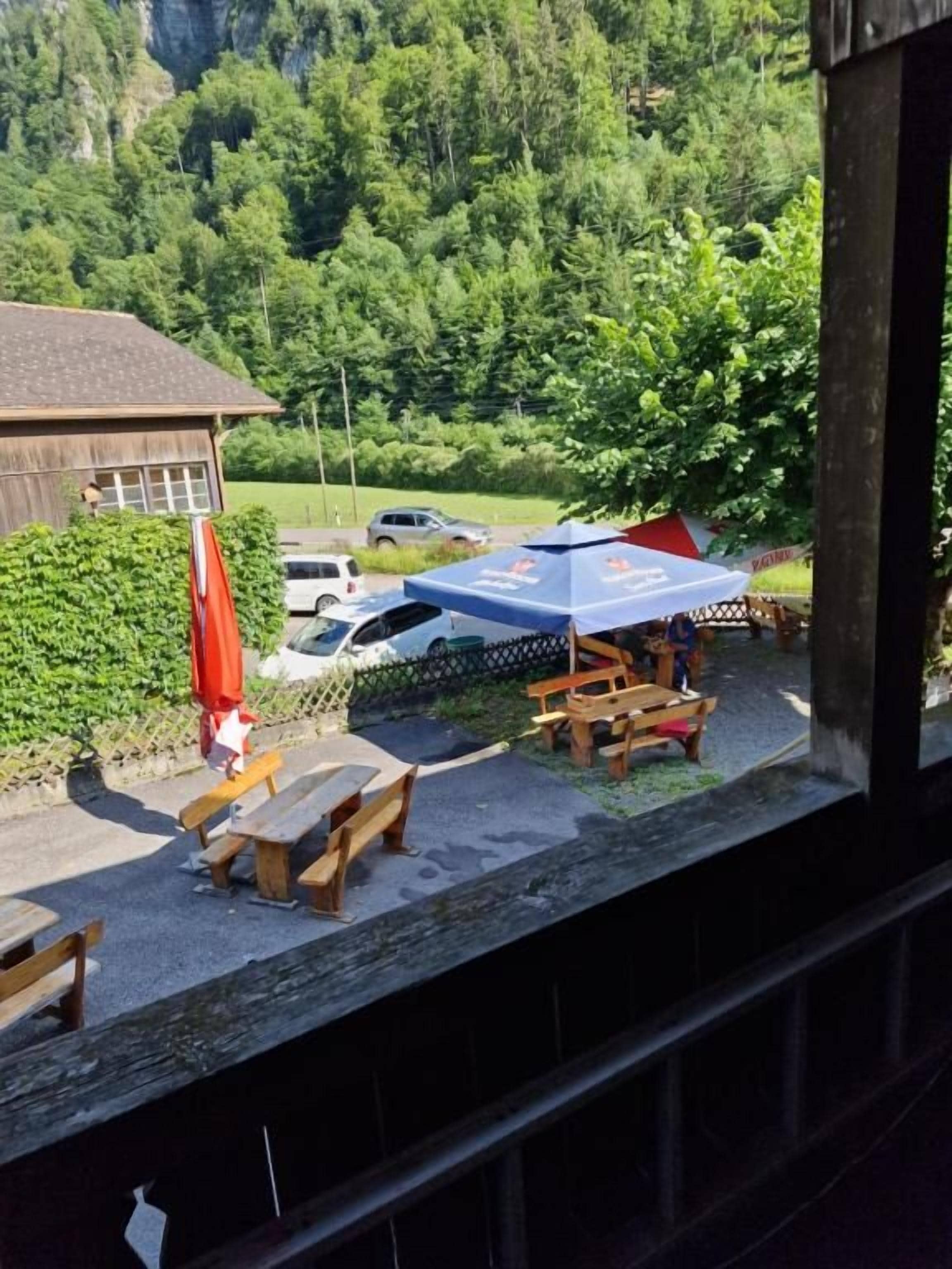 Una terraza de madera con mesas de picnic y un paraguas rojo, con vista a un estacionamiento y abundante vegetación.