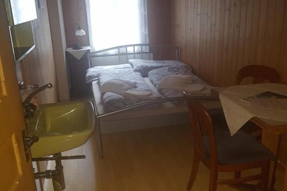 A small room with a bed, a table, a chair, a sink, and a window.