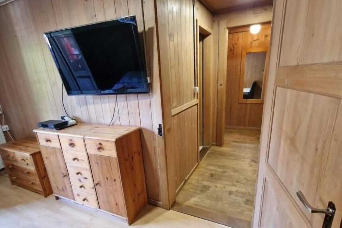 A room with wooden panel walls, a flat-screen TV mounted on the wall, a wooden cabinet with drawers, and a door leading to another room.