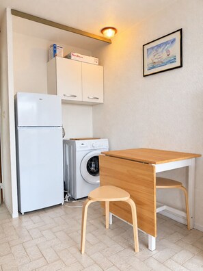 Een kleine keuken met een koelkast, een wasmachine en een houten tafel met een stoel.