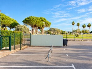 Een parkeerplaats met een groene omheining, een vuilnisbak en bomen op de achtergrond.