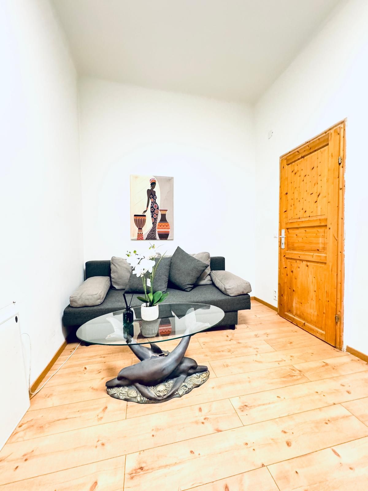 A modern living room with a glass coffee table, a sofa with cushions, a wooden door, and a framed artwork on the wall.