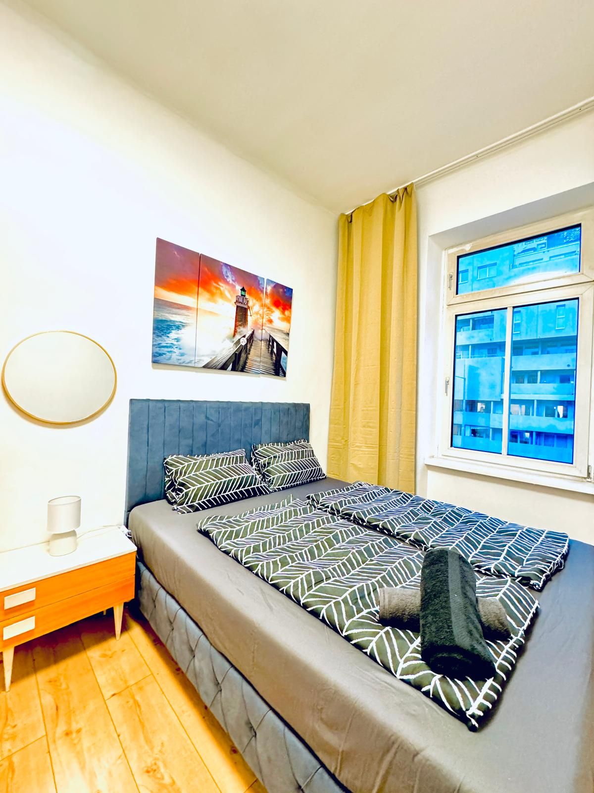 Een slaapkamer met een bed, nachtkastje, lamp, schilderij en raam met gordijnen.