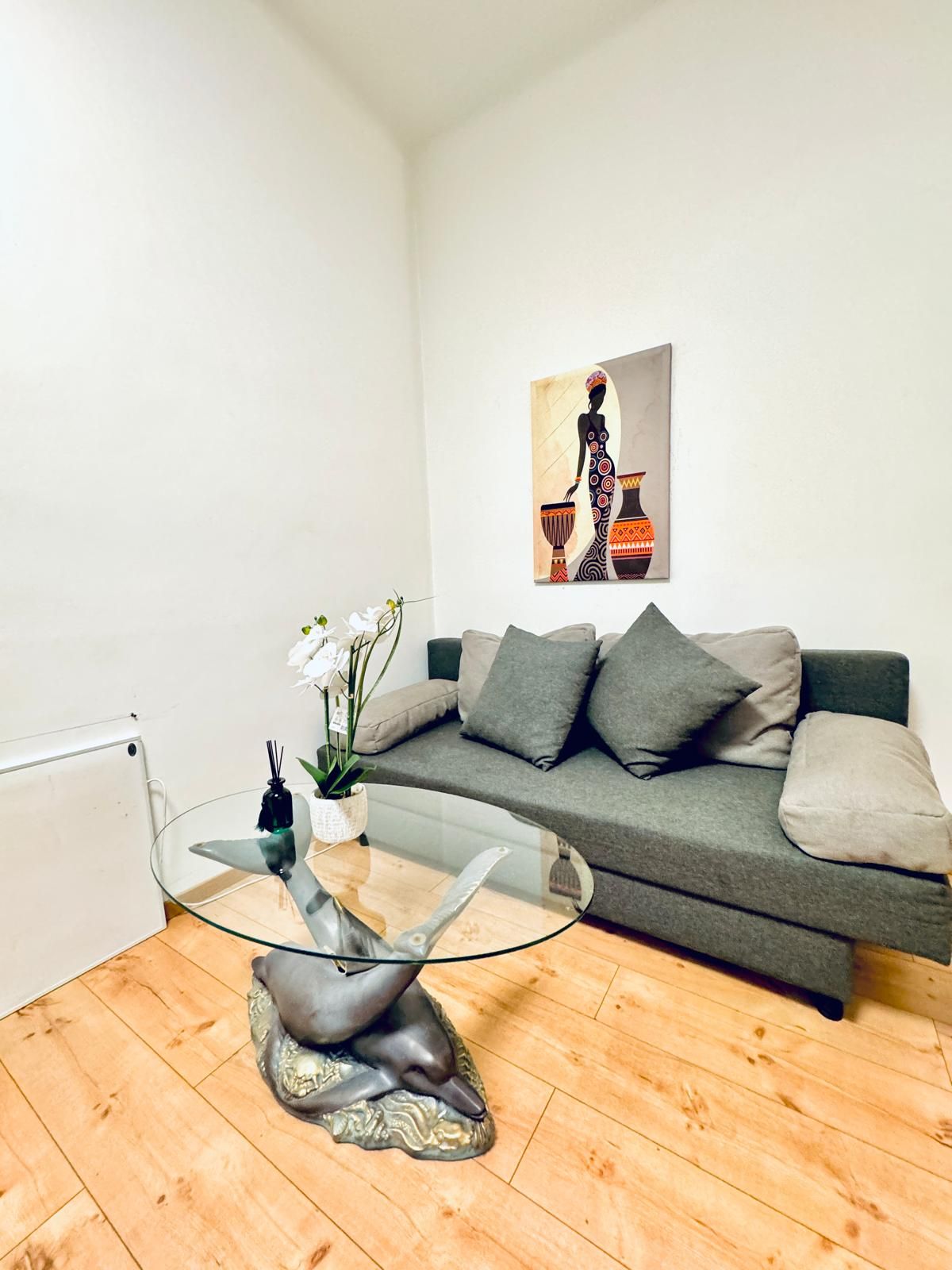 Een moderne woonkamer met een glazen salontafel, een grijze bank met kussens, een schilderij aan de muur en een potplant.