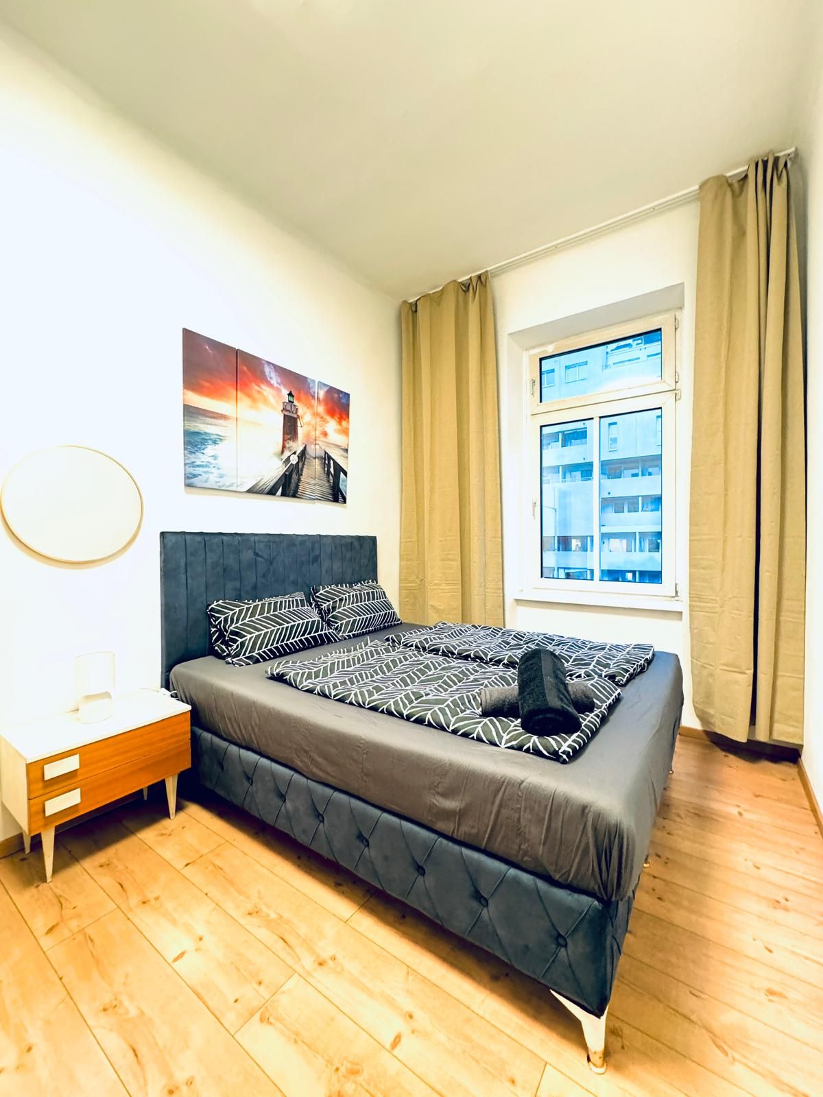 Een slaapkamer met een bed, nachtkastje, raam met gordijnen en een schilderij aan de muur.