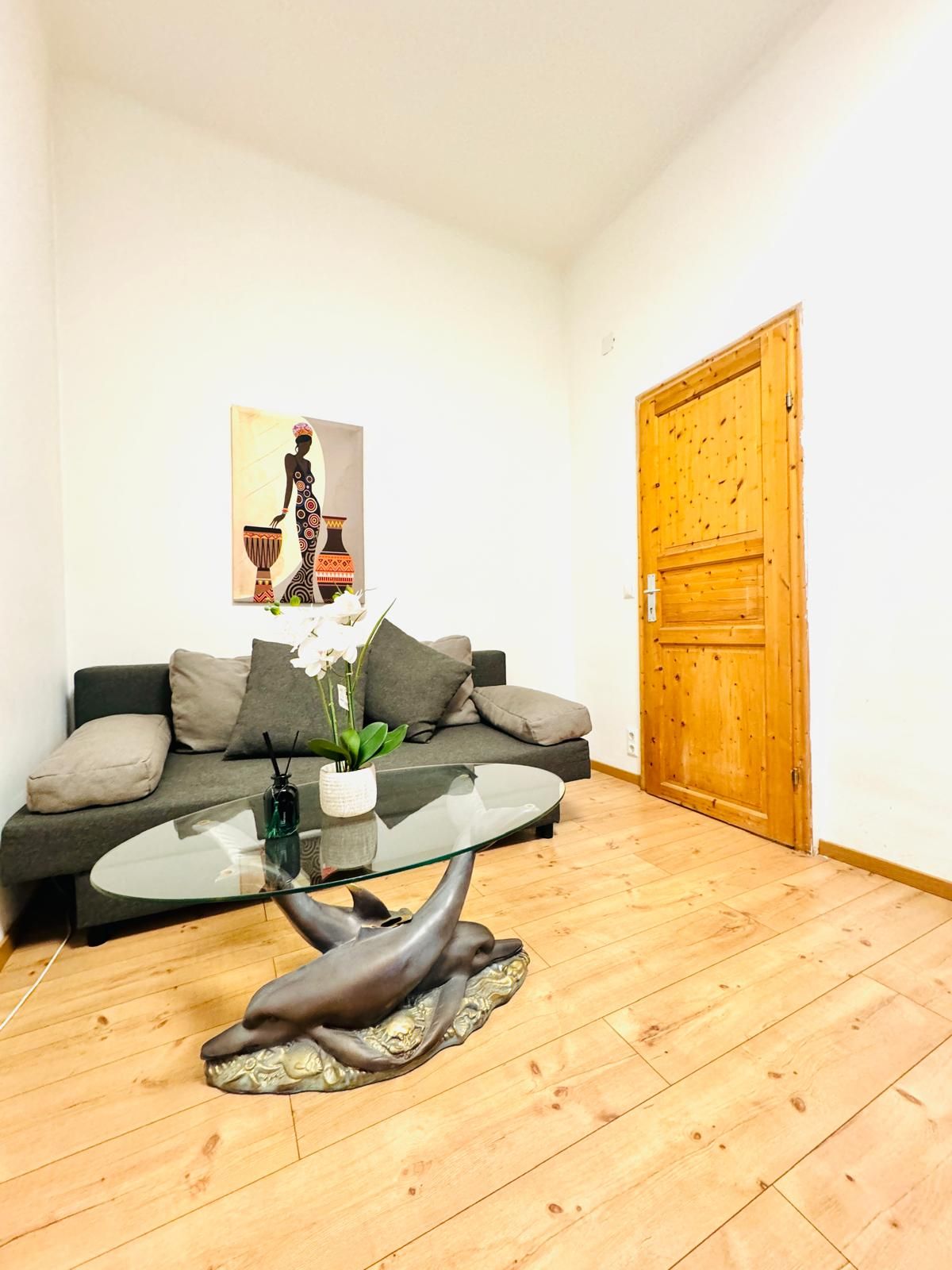 Een moderne woonkamer met een glazen salontafel, een bank met kussens, een houten deur en een schilderij aan de muur.