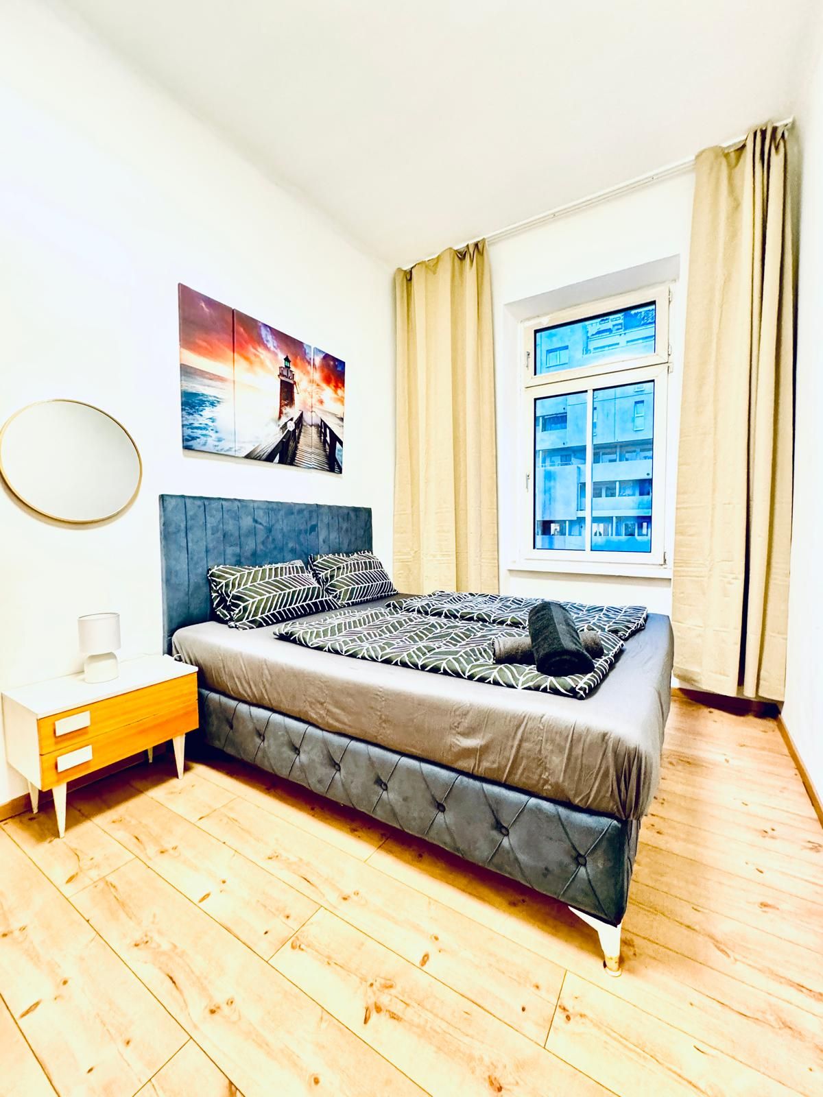 A bedroom with a bed, a bedside table, a window with curtains, and a wall-mounted picture.