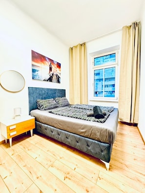 Un dormitorio con cama, mesita de noche, ventana con cortinas e imagen colgada en la pared.