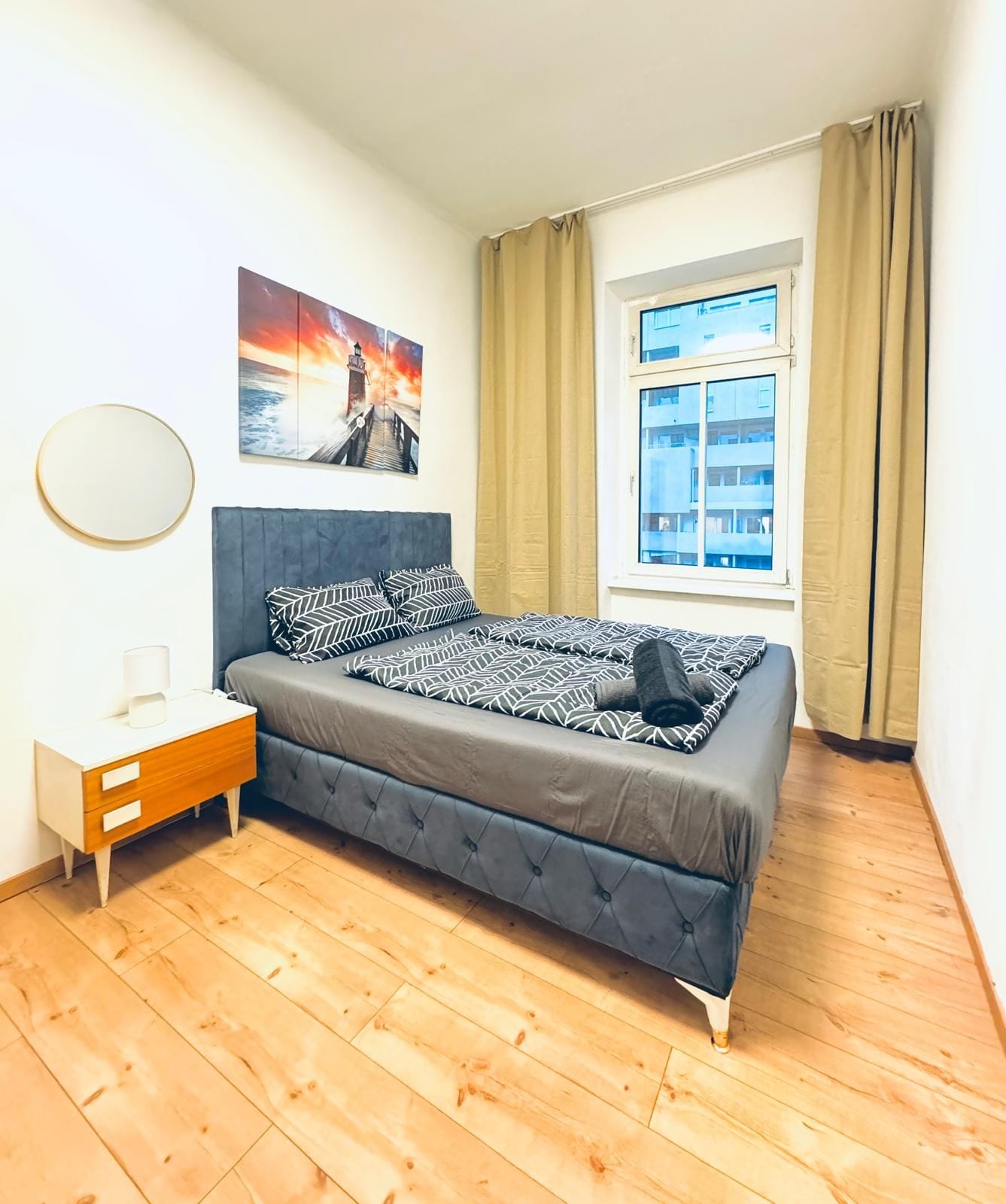 A bedroom with a bed, a bedside table, a window with curtains, a wall-mounted round mirror, and a painting.