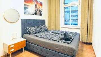 Ein Schlafzimmer mit Bett, Nachttisch, einem Fenster mit Vorhängen, einem runden Spiegel an der Wand und einem Gemälde.
