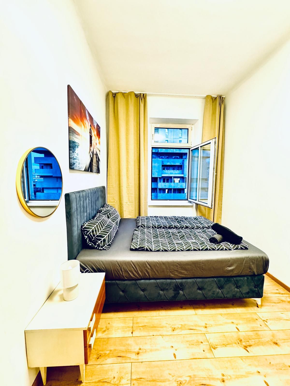 A bedroom with a bed, a bedside table, a mirror, a window with curtains, and a view of buildings outside.