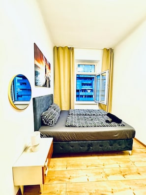 Un dormitorio con cama, mesita de noche, espejo, ventana con cortinas y vistas a edificios.