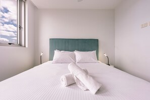 Een net opgemaakt bed met witte lakens en kussens, een blauwgroen hoofdbord en een raam met uitzicht op de lucht.
