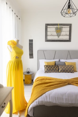 Une chambre à coucher avec un lit, une commode, un mannequin portant une robe jaune, un luminaire fixé au mur et un tableau encadré.