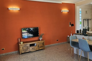 Habitació amb televisió penjada a una paret vermella, un armari de fusta, zona de menjador amb taula i cadires, i una finestra petita amb vistes.