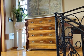 A wooden chest of drawers with multiple drawers, a white pedestal with a plant, and a staircase with ornate metal railings. - New Listing (Aprilia)