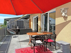 Uma varanda com mesa e cadeiras de madeira, um guarda-sol vermelho e uma estátua na parede.