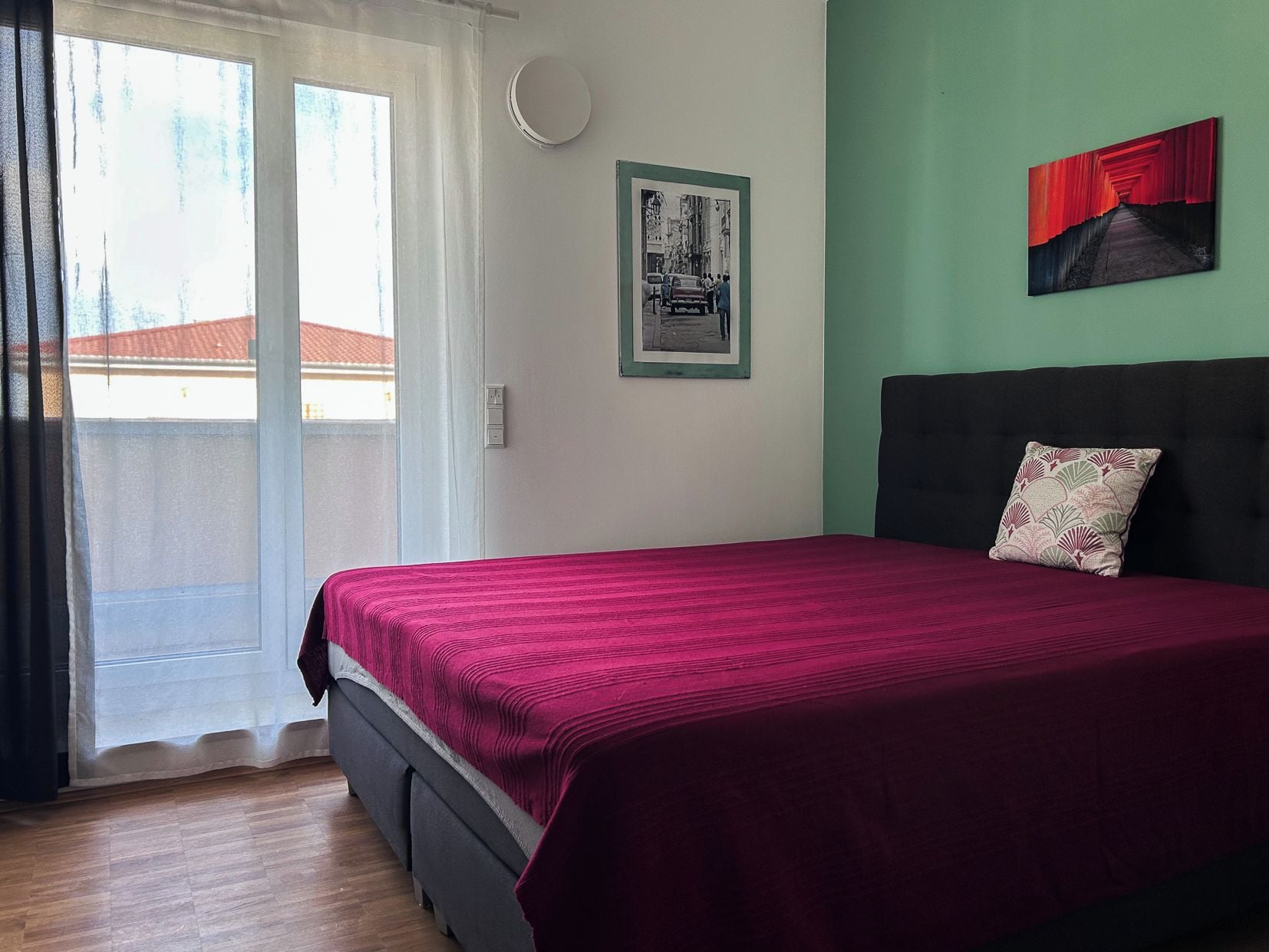 Un dormitorio con cama, ventana con cortinas, luz de pared e imagen enmarco.
