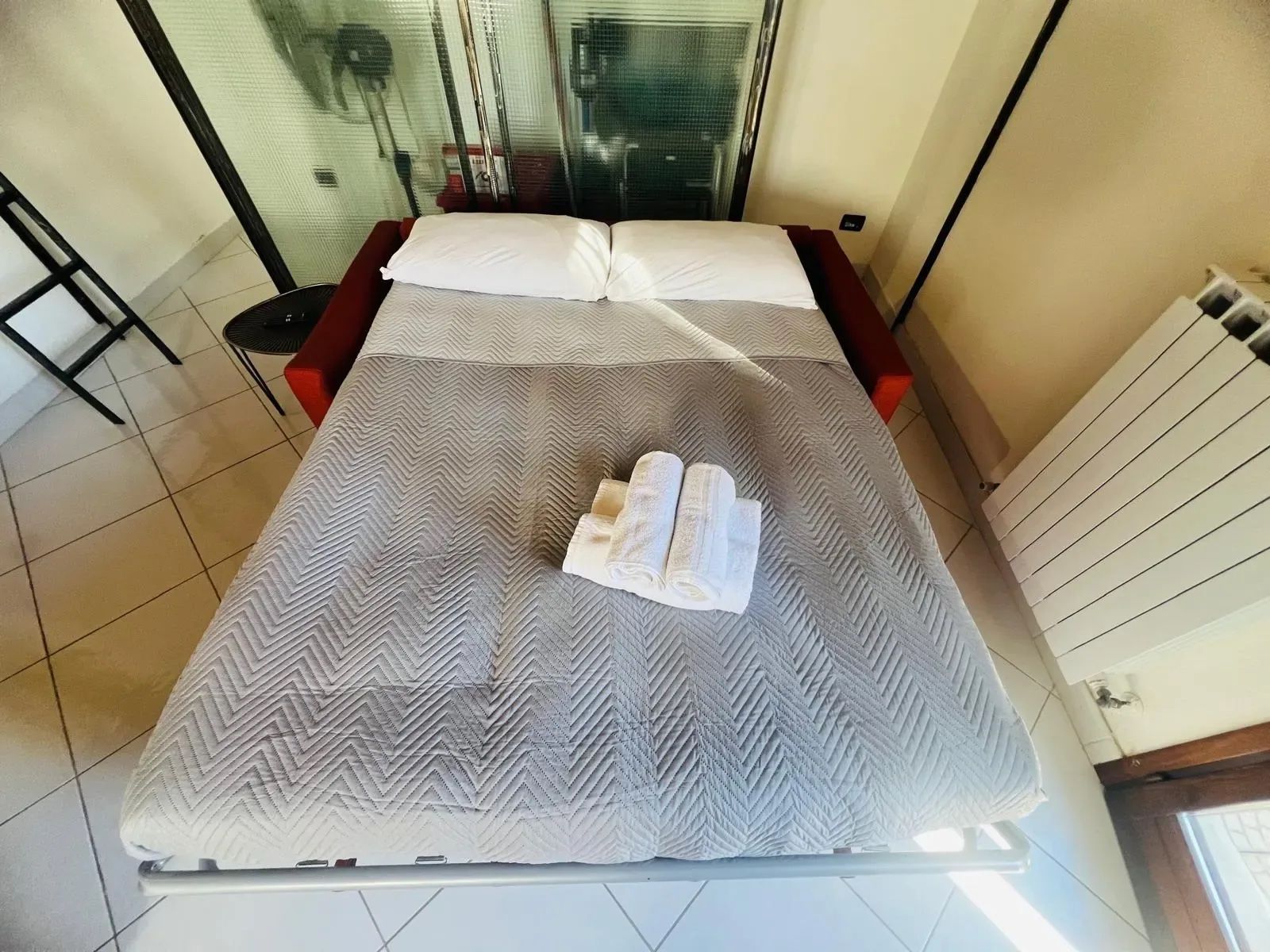 A bed with a patterned bedspread, two rolled towels, and a red bench beside it.