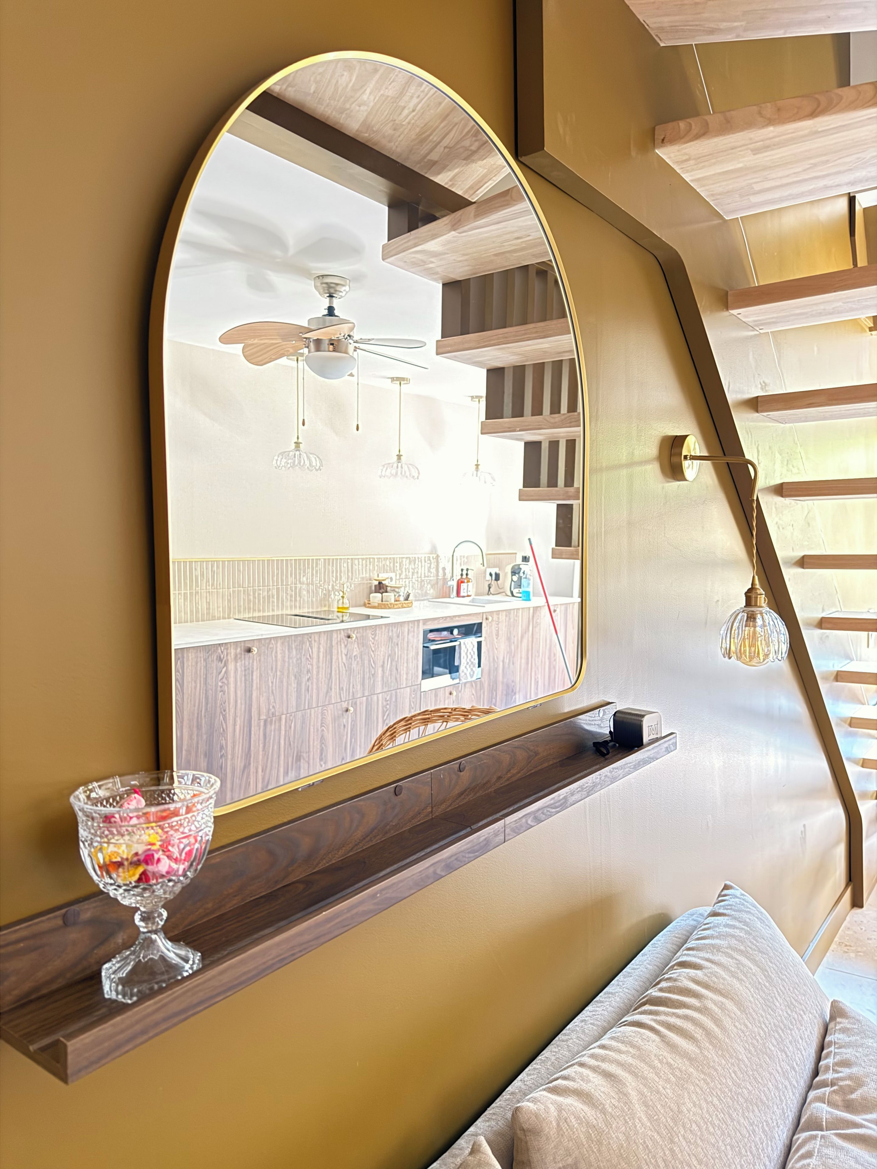 Un interior moderno con un espejo grande que refleja una zona de cocina, un estante de madera con un cuenco de vidrio y una escalera de diseño único.