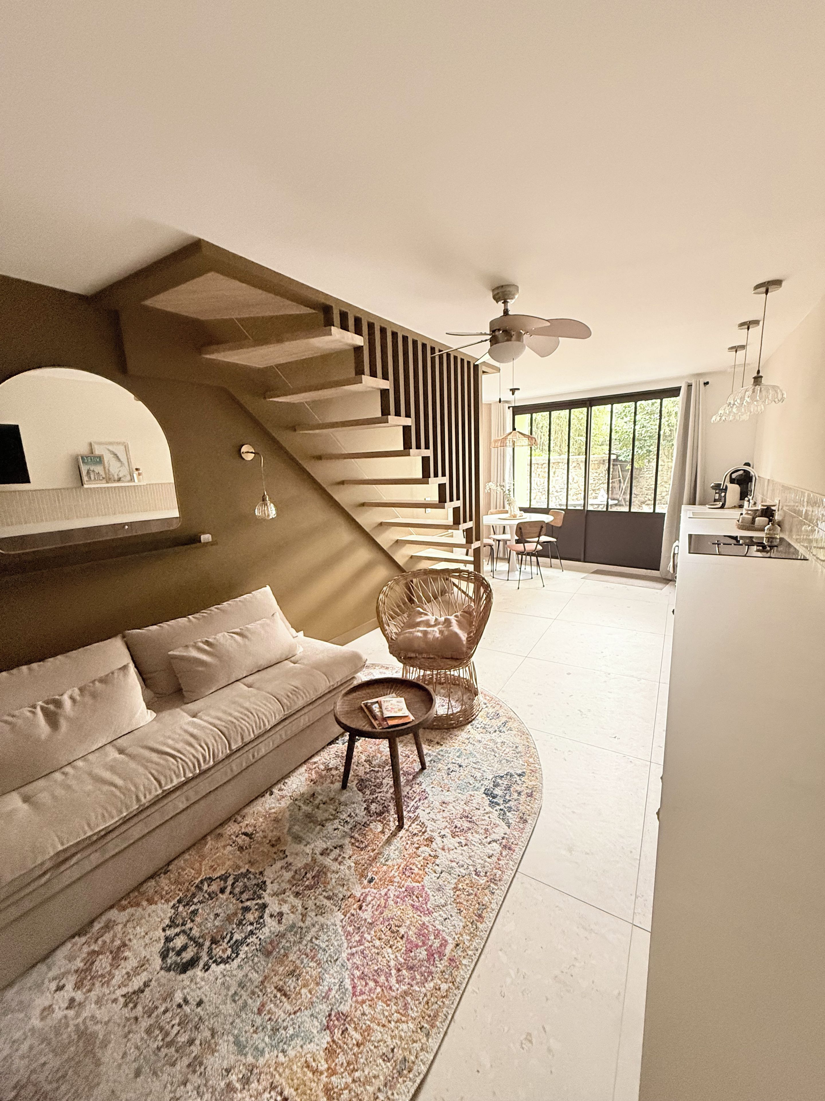 Una sala de estar moderna con un sofá, una mesa redonda para café, una silla de mimbre, una escalera y vistas al exterior.