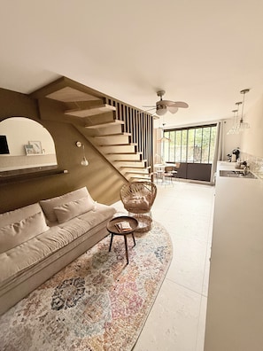 Una sala de estar moderna con un sofá, una mesa redonda para café, una silla de mimbre, una escalera y vistas al exterior.