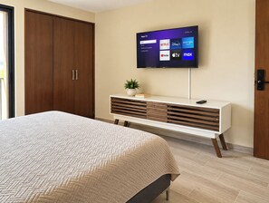 Un dormitorio con cama, televisor montado en la pared, armario de madera y mesita de noche con una planta.