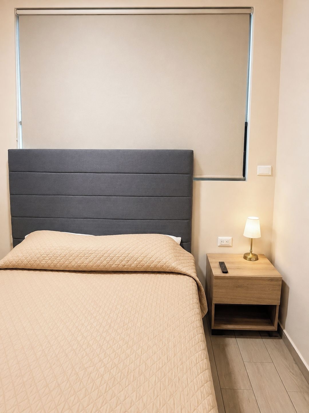 A bed with a quilted beige cover, a dark headboard, and a bedside table with a lamp.