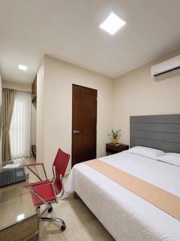 Une chambre d’hôtel avec un lit, un bureau avec un ordinateur portable, une chaise rouge, une table de chevet avec une plante et un appareil de climatisation.