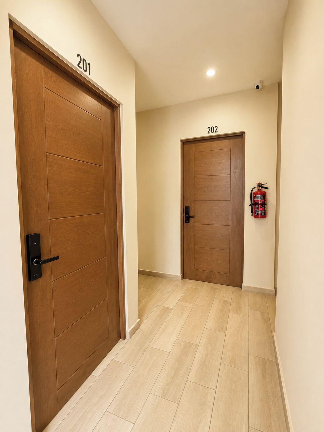 A hallway with two wooden doors labeled 201 and 202, a fire extinguisher mounted on the wall, and a ceiling-mounted security camera.