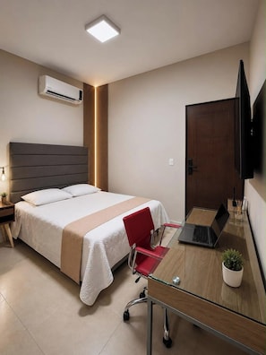 Une chambre d’hôtel avec un lit, un bureau avec un ordinateur portable, une chaise rouge, un téléviseur fixé au mur et un appareil de climatisation.