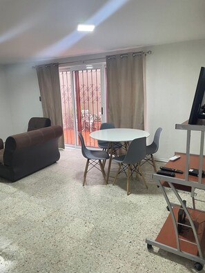 Ein Zimmer mit einem runden Tisch, vier Stühlen, einer Couch, einem Fernseher auf einem Schrank und einer Glastür zum Außenbereich.