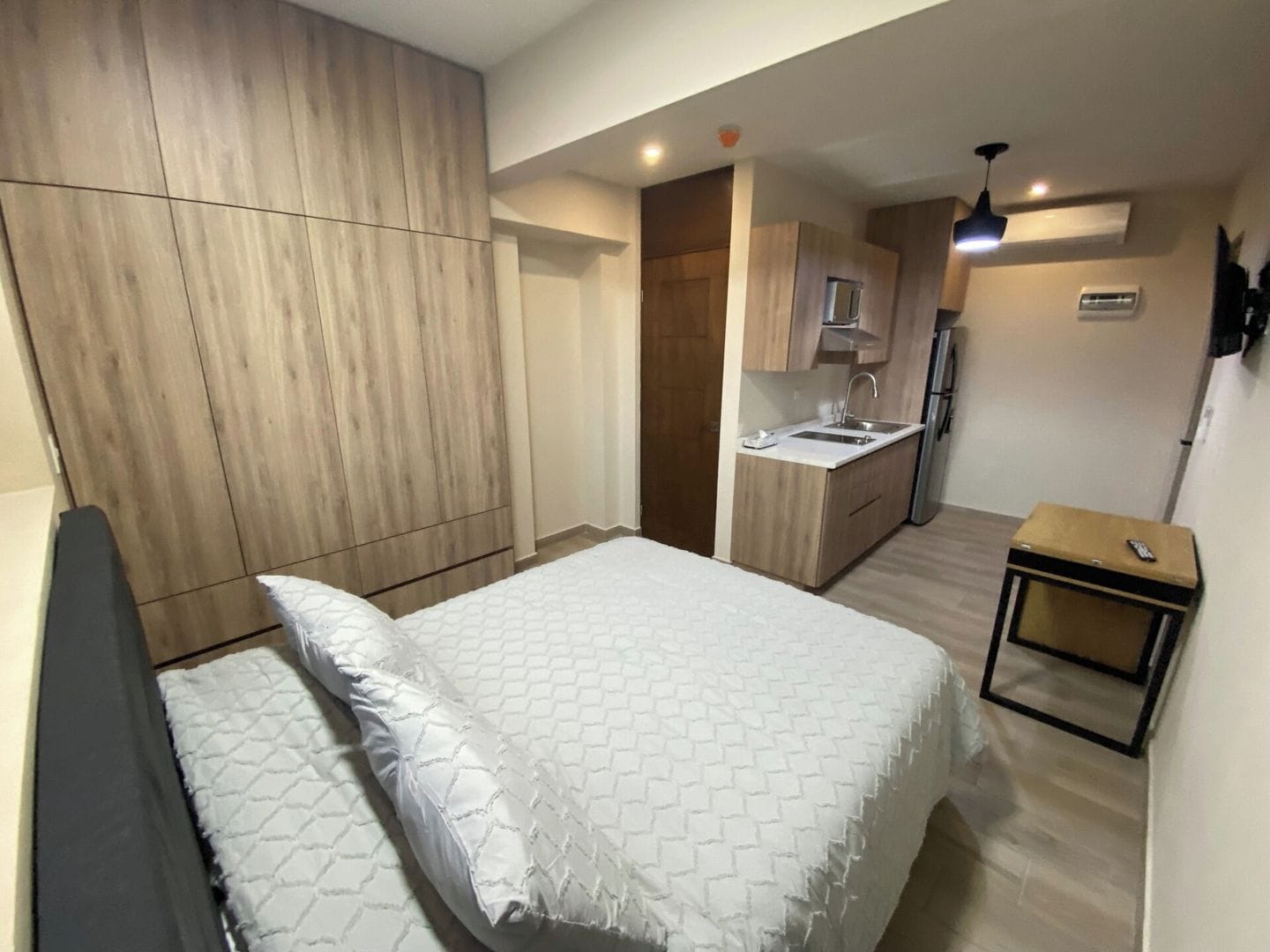 A bedroom with a bed, a bedside table, and a small kitchenette with a sink and a refrigerator.