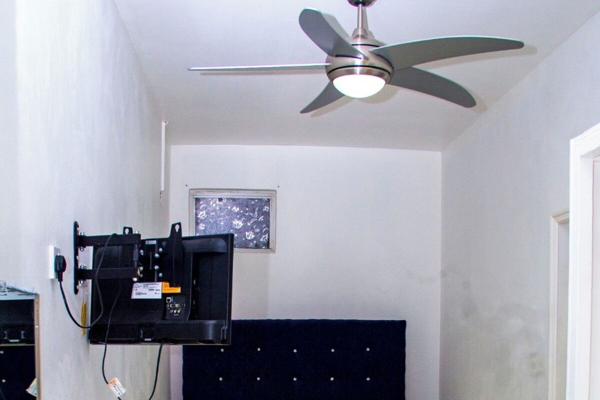 A hotel room with a bed, a ceiling fan, a mounted television, and a bedside table with a lamp.