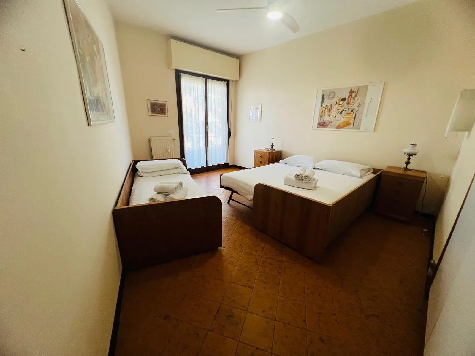 Ein Hotelzimmer mit zwei Betten, einem kleinen Tisch, einem Nachttisch, einem Heizkörper und einem Fenster mit Vorhängen.