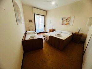 A hotel room with two beds, a small table, a nightstand, a radiator, and a window with curtains. - New Listing (Sanremo)