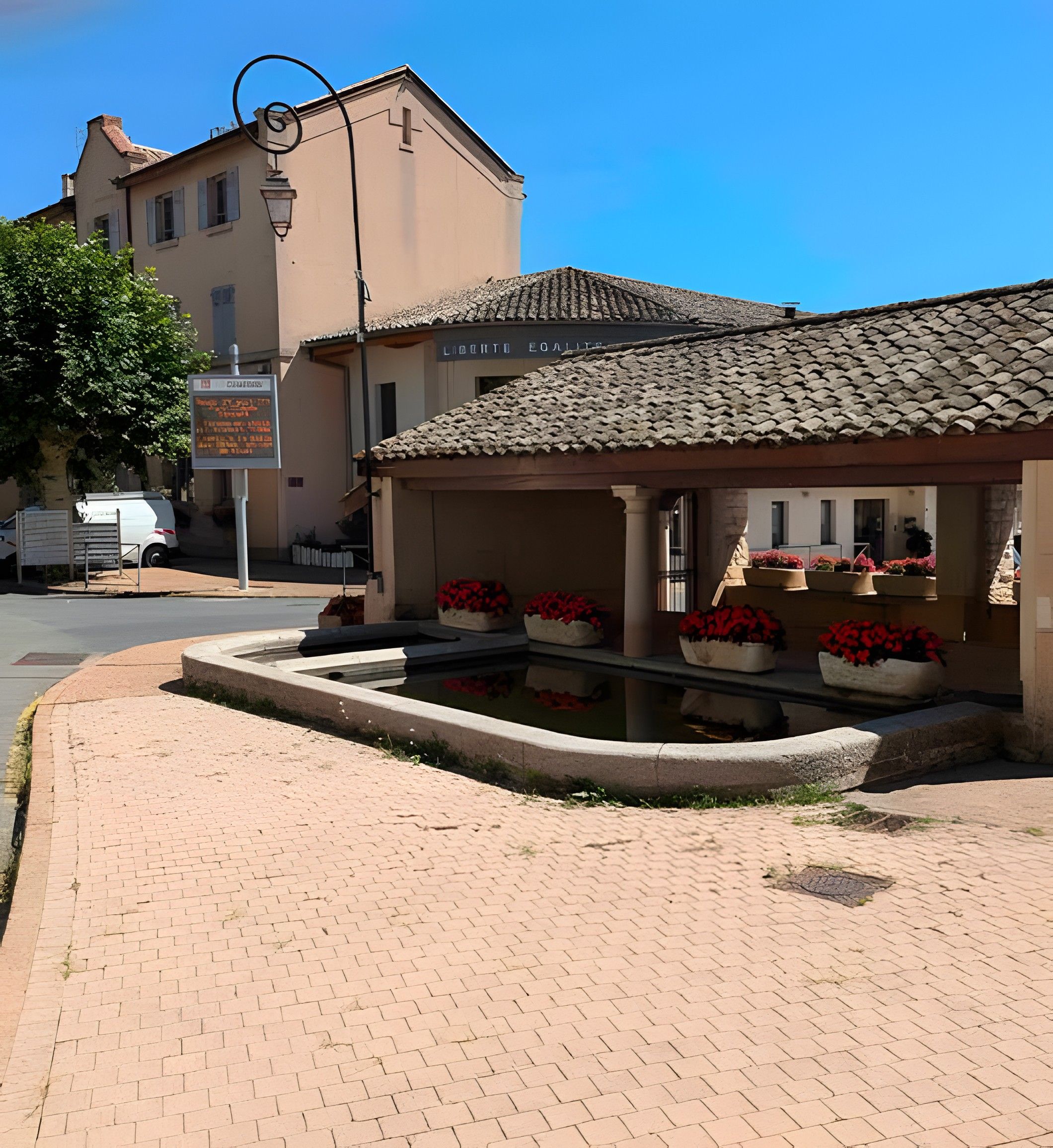 A building with a tiled roof, a paved courtyard with flower planters, and a sign with text.