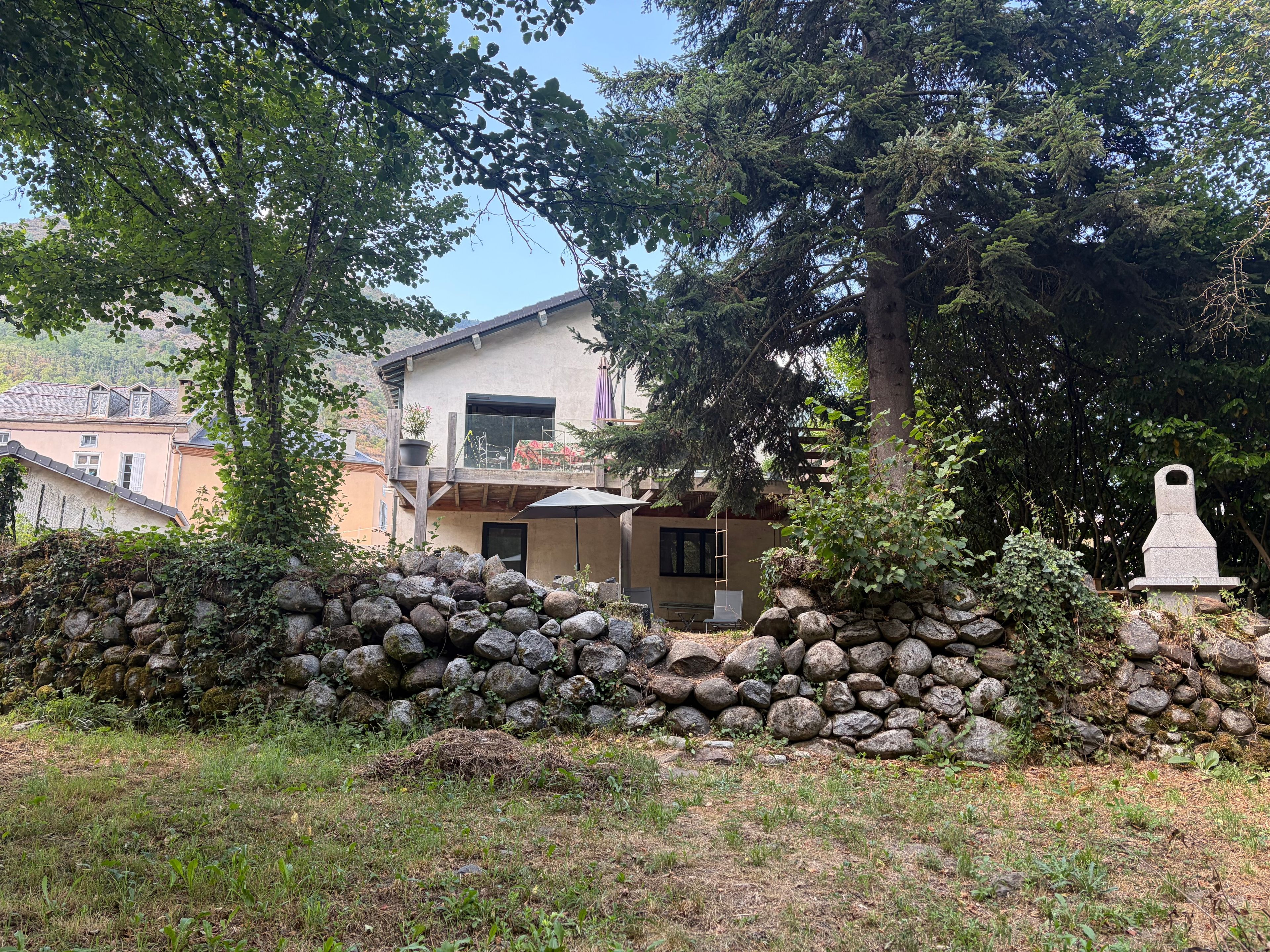 Un mur de pierre bordant un petit jardin, une terrasse en bois et un bâtiment doté d’un toit en pente.