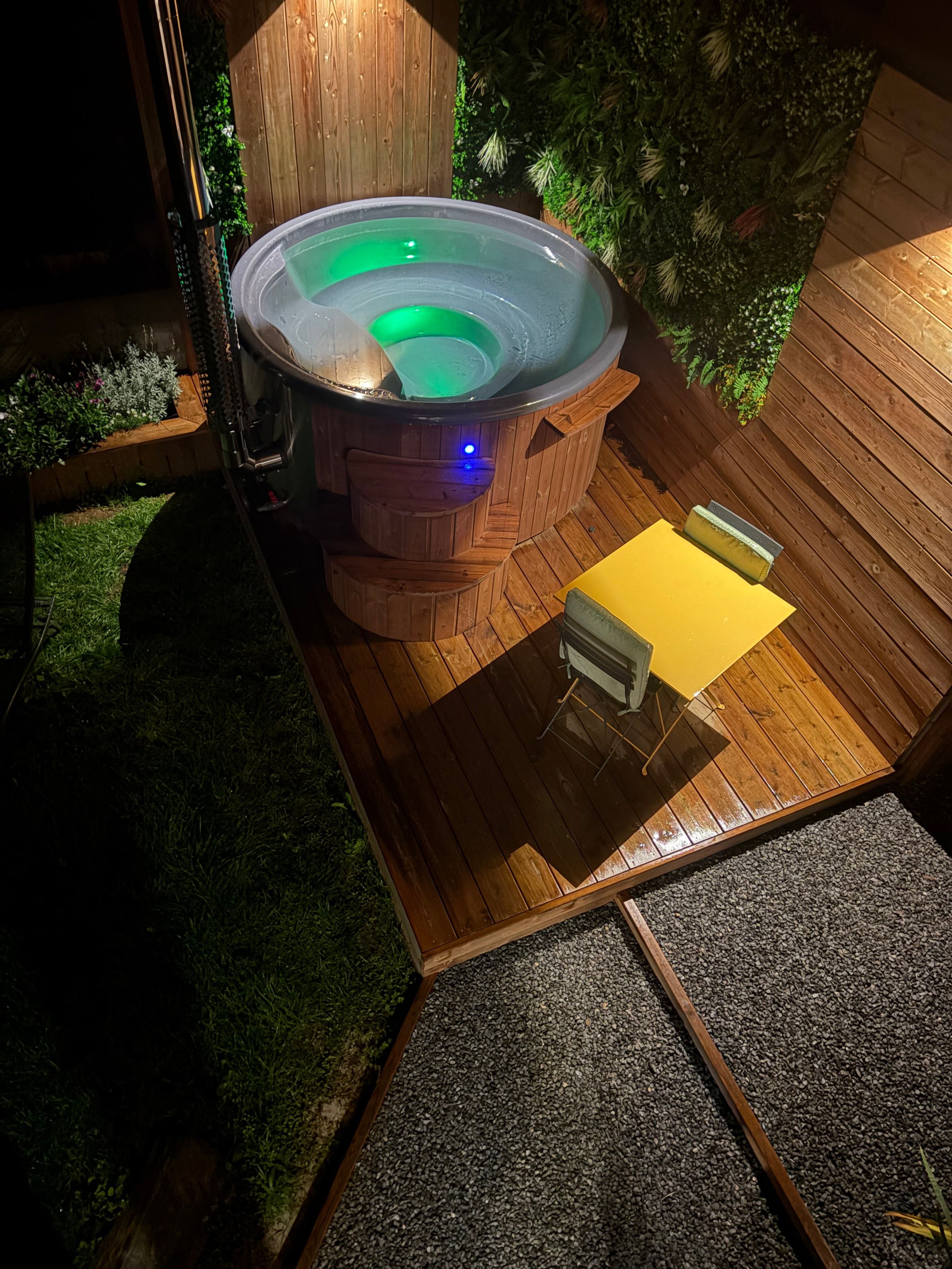 A hot tub with a wooden deck and a small table, surrounded by a wooden fence and greenery.